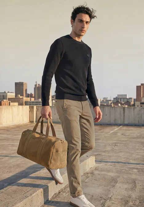 Ein Mann läuft auf einem Dach mit einer Reisetasche, trägt schwarzen Pullover und beige Hose.