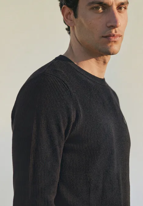 Ein Mann in schwarzem Pullover blickt seitlich in die Kamera vor neutralem hellem Hintergrund.