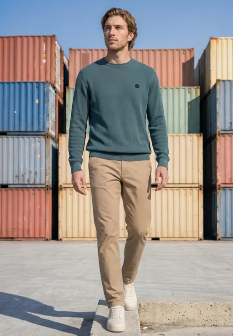 Ein Mann mit grünem Pullover und beigen Hosen spaziert vor bunten Frachtcontainern in einem Industriehafen.