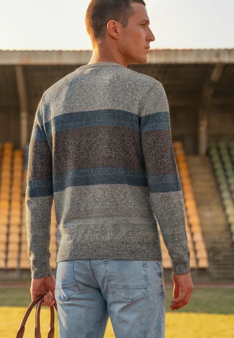 Ein Mann in gestreiftem Pullover und Jeans steht von hinten auf einem Sportfeld vor leeren Tribünen.