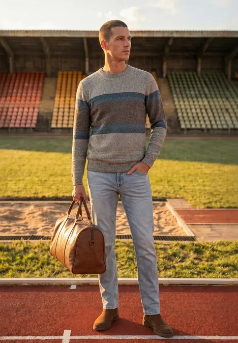 Ein Mann im gestreiften Pullover mit Reisetasche steht auf der Tartanbahn vor bunten Tribünen.