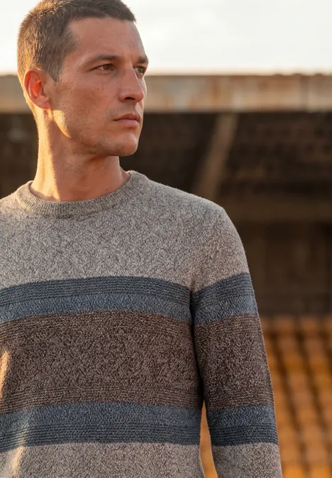 Ein Mann im gestreiften Pullover blickt nach rechts vor einer Tribüne im Freien.