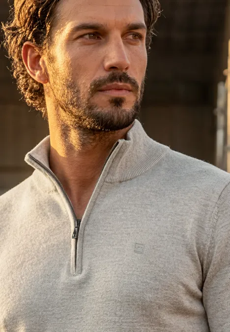 Een man met baard en donker krullend haar draagt een lichtgrijze trui met halve rits en kijkt opzij.