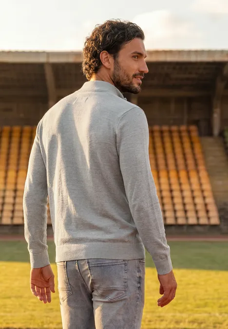 Een man in een grijze trui en spijkerbroek staat op een sportveld en kijkt over zijn schouder.