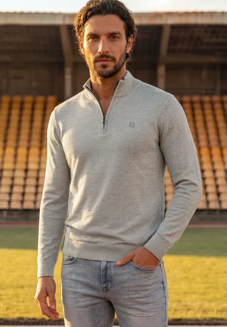Een man in een grijze trui en spijkerbroek staat voor lege tribunes in een stadion.