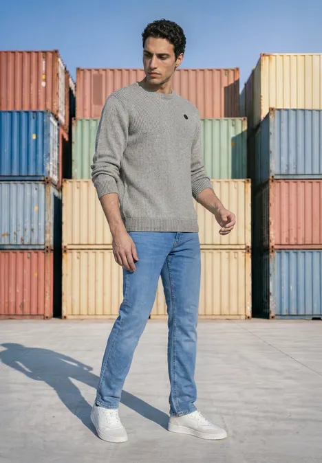 Mouliné Pullover mit Rundhals silver grey melange Mann in grauem Pullover, blauen Jeans und weißen Sneakern vor pastellfarbenen Frachtcontainern.