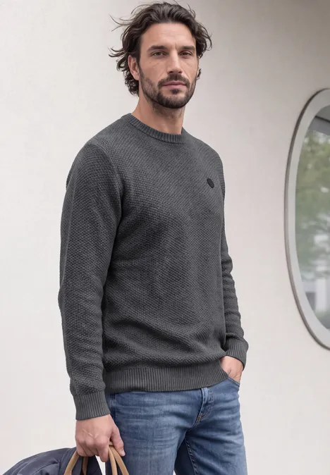 Sweter o wyglądzie melanżowym z strukturą light anthra grey melangey Mężczyzna w szarym swetrze i dżinsach stoi przed ścianą z okrągłym oknem.