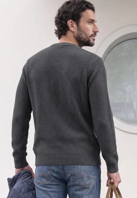 Sweter o wyglądzie melanżowym z strukturą light anthra grey melangey Mężczyzna w szarym swetrze widoczny z profilu przed okrągłym oknem.