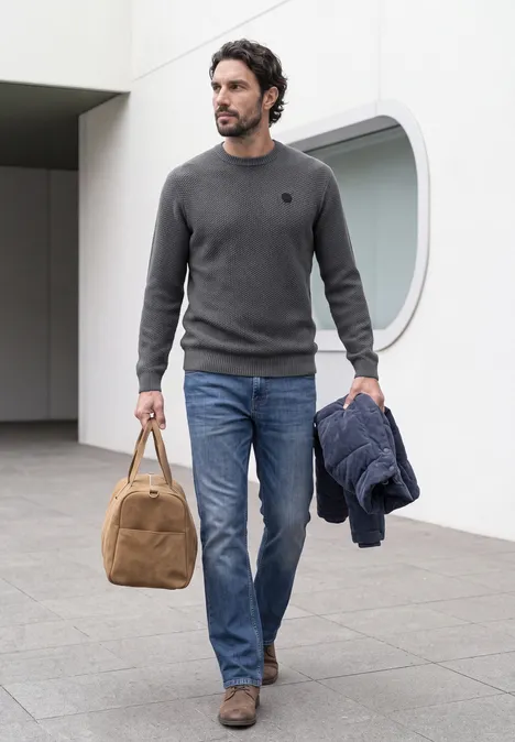Sweter o wyglądzie melanżowym z strukturą light anthra grey melangey Mężczyzna w dżinsach i swetrze niesie torbę i trzyma kurtkę.