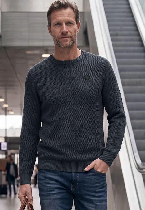 Sweter o strukturze melanżowej dark basic blue melange Mężczyzna w dżinsach i swetrze stoi w przestrzeni publicznej obok ruchomych schodów.