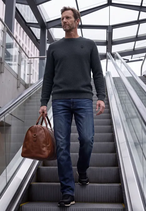 Sweter o strukturze melanżowej dark basic blue melange Mężczyzna w dżinsach i swetrze niesie brązową skórzaną torbę na schodach ruchomych w nowoczesnym przeszklonym budynku.
