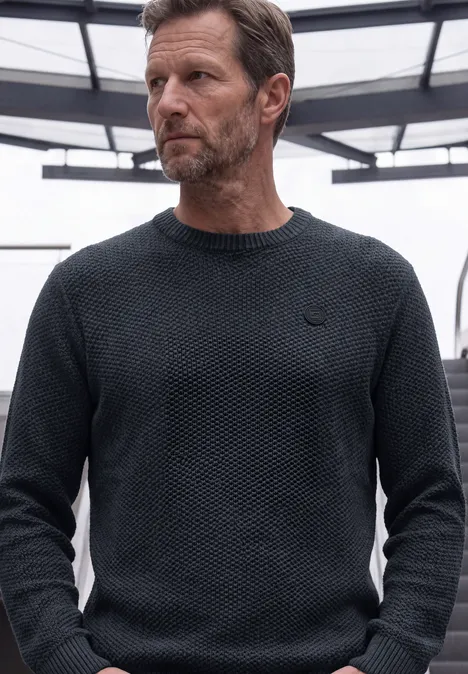 Sweter o strukturze melanżowej dark basic blue melange Mężczyzna w ciemnym, dzianym swetrze spogląda w lewo na tle nowoczesnej architektury.