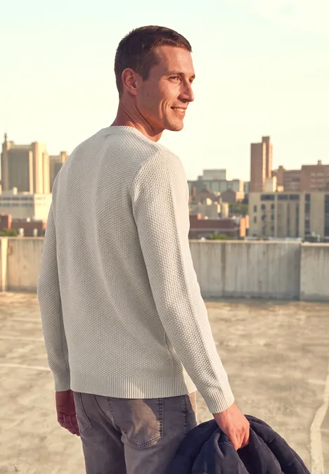 Sweter w stylu melanżowym z strukturą bright beige melange Mężczyzna w jasnym swetrze trzyma kurtkę i podziwia miejską panoramę z dachu.