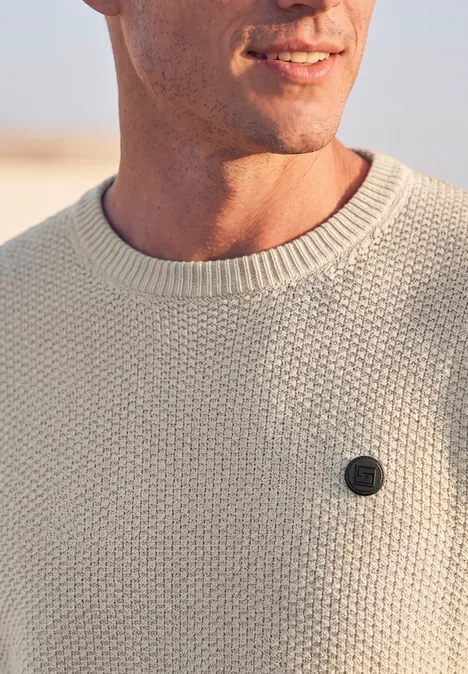 Sweter w stylu melanżowym z strukturą bright beige melange Zbliżenie na mężczyznę w kremowym swetrze z drobnym okrągłym logo na wysokości piersi.