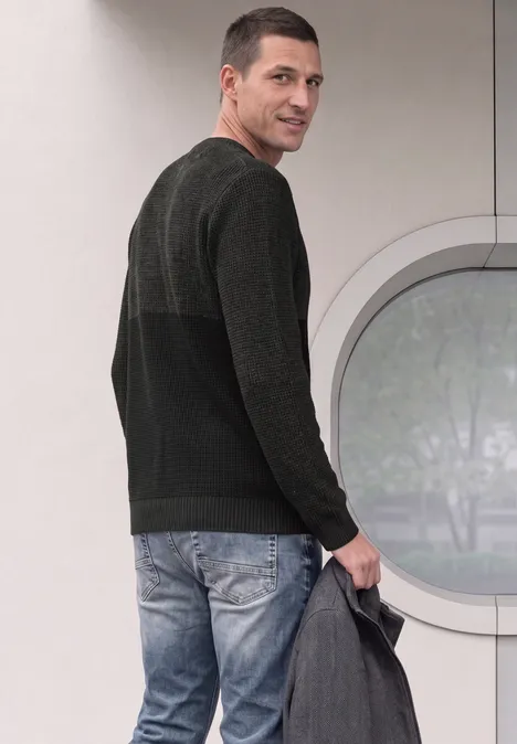 Sweter z okrągłym dekoltem i elementami w stylu mouliné anthra grey melange Mężczyzna w dżinsach i ciemnym swetrze spogląda przez ramię, trzymając szarą kurtkę.