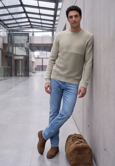 Sweter z okrągłym dekoltem i elementami w stylu mouliné bright beige melange W luźnym ubraniu mężczyzna opiera się o ścianę w jasnym atrium, obok stoi duża torba podróżna.