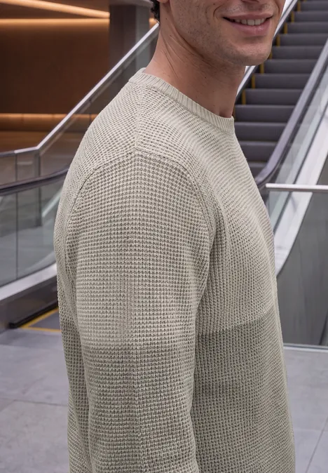 Sweter z okrągłym dekoltem i elementami w stylu mouliné bright beige melange Uśmiechnięty mężczyzna w dwukolorowym swetrze przed ruchomymi schodami w nowoczesnym otoczeniu.
