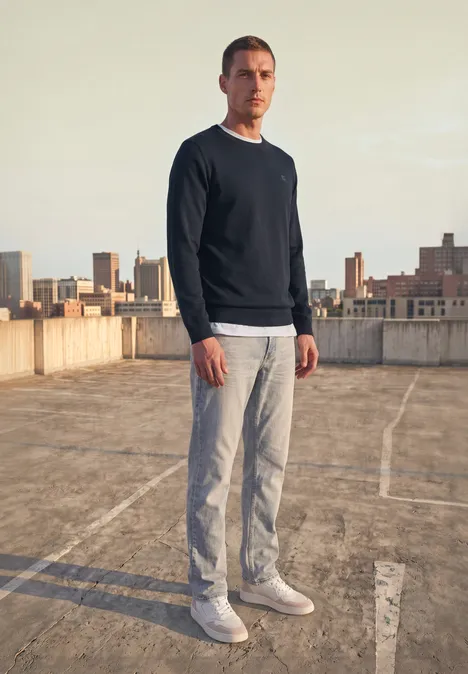 Ein Mann in Pullover und Hose steht auf einem Dachparkplatz mit Stadtkulisse im Hintergrund.