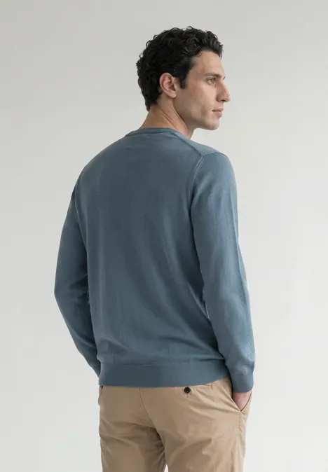 Ein Mann im blau-grauen Pullover und beiger Hose steht seitlich, eine Hand in der Tasche.