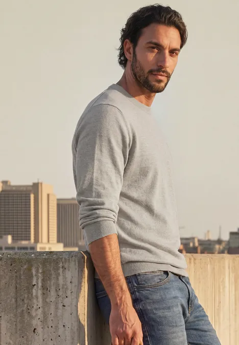 Ein lässig gekleideter Mann lehnt sich vor einer Betonmauer ab, trägt grauen Pullover und Jeans.