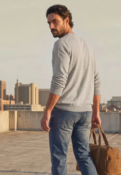 Mann steht auf einem Dach, blickt zurück, trägt einen grauen Pullover, Jeans und hält eine Reisetasche.
