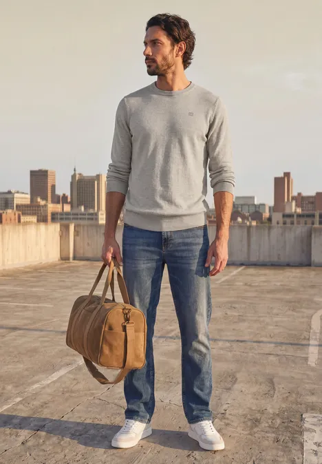 Mann im Pullover, Jeans und Sneakers, trägt Reisetasche auf Dach mit Stadtblick.