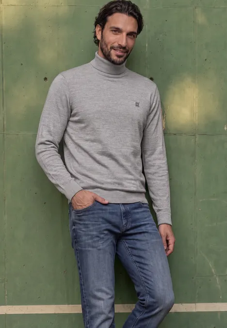 Basic sweter z golfem silver grey melange Mężczyzna w szarym golfie i dżinsach stoi swobodnie przed zieloną ścianą.