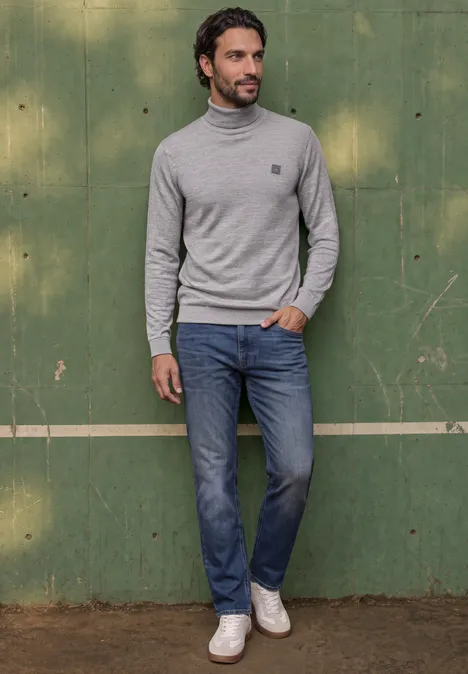 Basic sweter z golfem silver grey melange Mężczyzna w szarym golfie, niebieskich dżinsach i białych trampkach na tle zielonej drewnianej ściany.