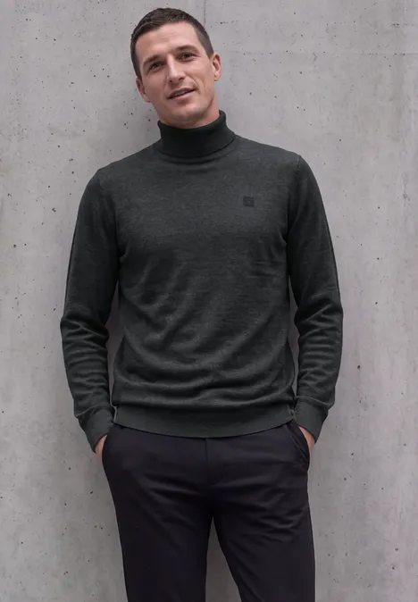Basic sweter z golfem anthra grey melange Mężczyzna stoi przed szarą ścianą z rękami w kieszeniach, ubrany w ciemny golf.