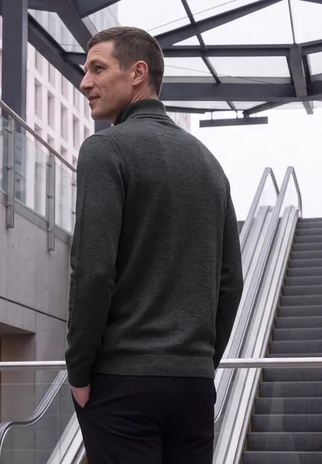 Basic sweter z golfem anthra grey melange Mężczyzna w szarym swetrze stoi przy ruchomych schodach we współczesnym budynku.