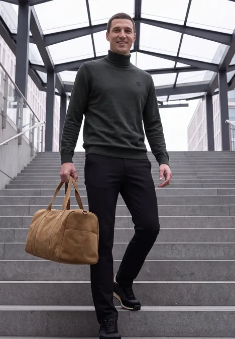 Basic sweter z golfem anthra grey melange Uśmiechnięty mężczyzna schodzi po schodach w nowoczesnym szklanym przejściu, niosąc brązową torbę podróżną.