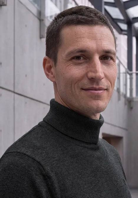 Basic sweter z golfem anthra grey melange Mężczyzna z krótkimi brązowymi włosami w ciemnym golfie stoi w nowoczesnym korytarzu.