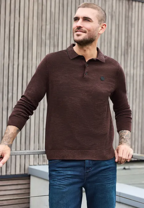 Mann trägt braunen Pullover mit Knopfleiste und blaue Jeans, steht lässig vor Holzlamellen