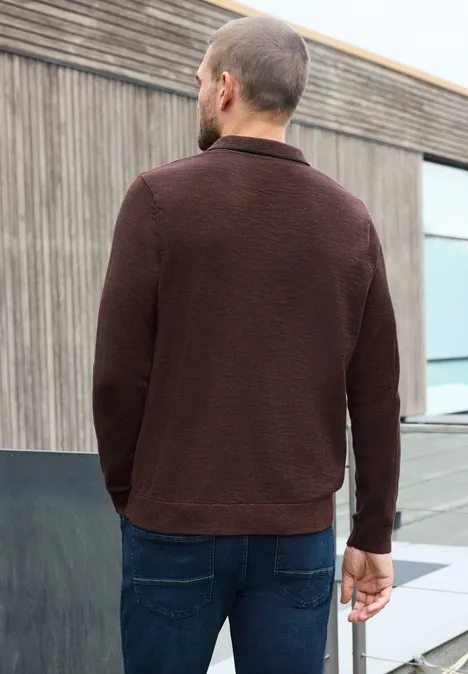 Ein Mann in braunem Pullover und Jeans steht vor einem modernen Gebäude mit Holzverkleidung.