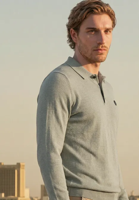 Een man met bruin haar draagt een grijze polo met lange mouwen en staat voor een stadsgezicht.
