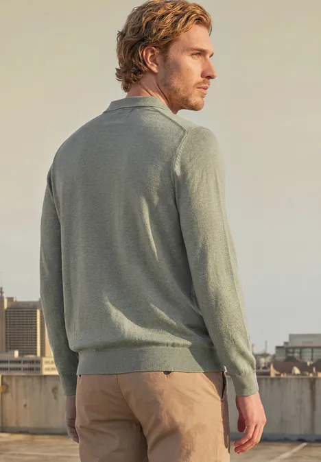 Ein Mann im grauen Pullover und beigen Hosen steht auf einem Dach, schaut über die Stadt.