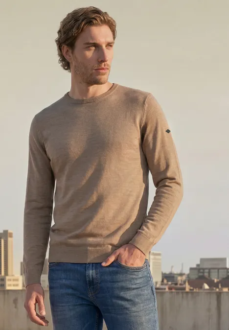 Ein junger Mann in braunem Pullover und Jeans steht auf einem Dach mit urbaner Skyline im Hintergrund.
