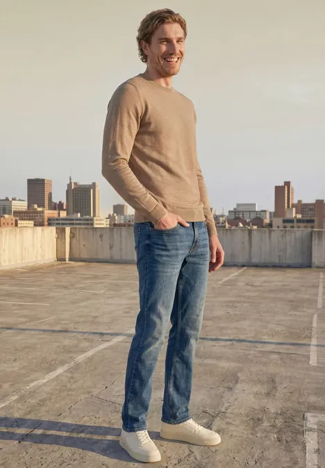 Ein lächelnder Mann in Jeans und beigem Pullover steht auf einem Parkdeckdach mit Stadtblick.