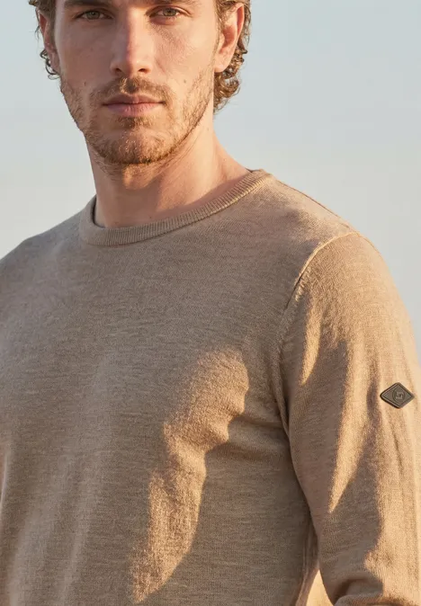 Ein Mann mit Bart trägt einen hellbeigen Pullover vor neutralem Hintergrund.