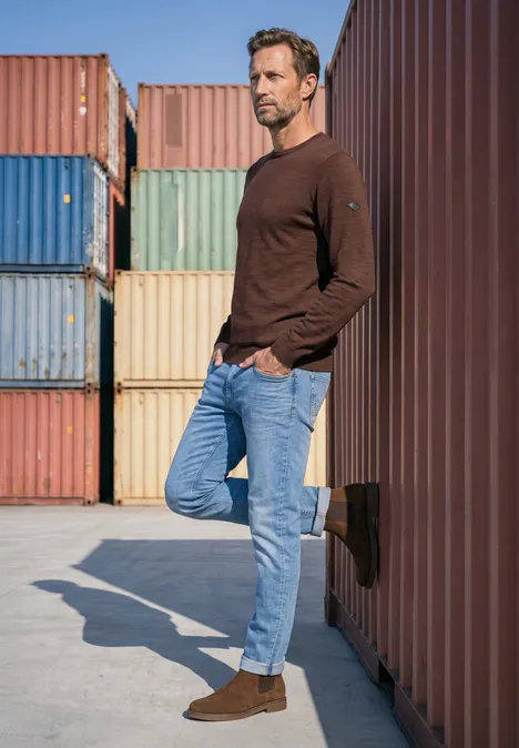 Ein lässig gekleideter Mann steht vor bunten Seecontainern und lehnt entspannt gegen einen Container.