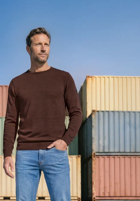 Ein Mann in braunem Pullover und Jeans steht vor bunten Seecontainern.