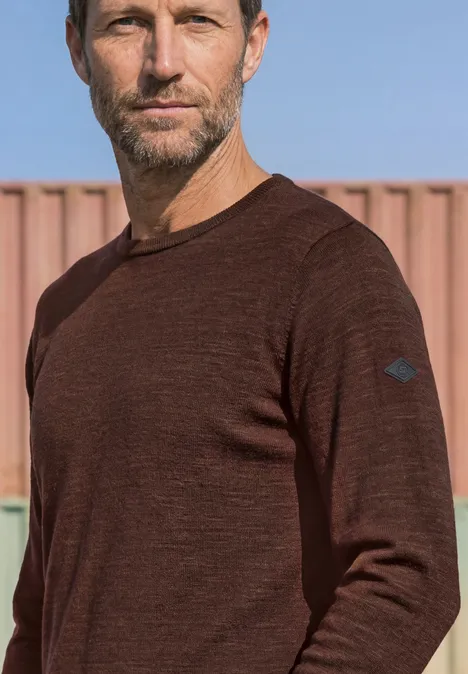Ein Mann mit kurzem Haar und Bart trägt ein braunes, langärmeliges Shirt vor einem Containerhintergrund.