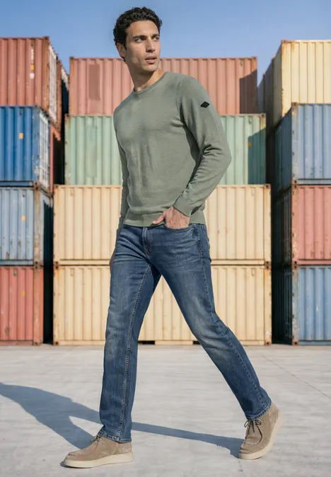 Ein Mann trägt grünes Langarmshirt, Jeans und helle Schuhe, steht vor bunten Frachtcontainern.