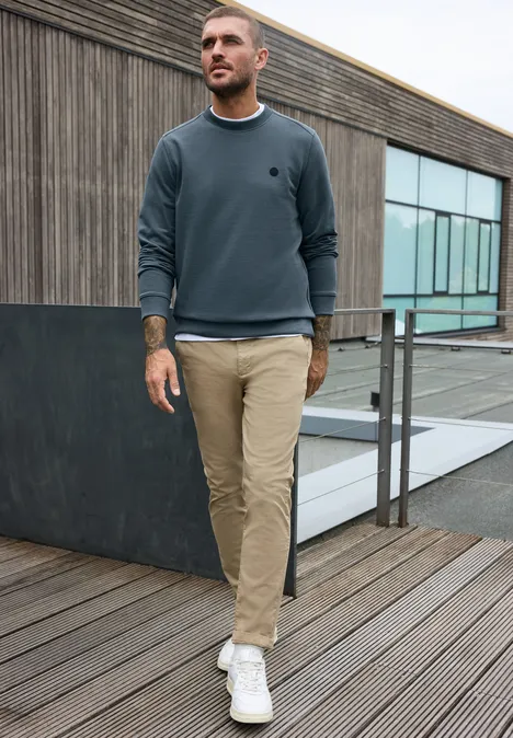 Ein junger Mann in grauem Sweatshirt und beigen Hosen spaziert auf einer Holzterrasse vor modernem Gebäude.