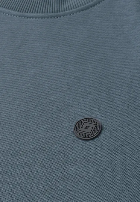 Nahaufnahme eines blauen Sweatshirts mit rundem schwarzen Logo-Patch neben dem Kragen.