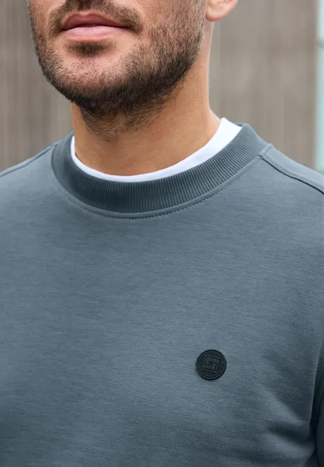 Ein Mann in einem grauen Sweatshirt mit rundem, schwarzen Logo auf der Brust steht vor einem verschwommenen Hintergrund.