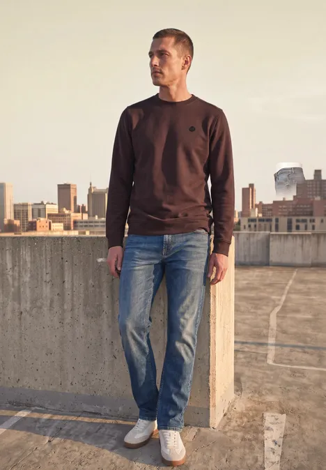 Ein Mann steht auf einem Parkdeck und blickt auf die Stadt. Er trägt braunes Sweatshirt, Jeans und weiße Sneaker.
