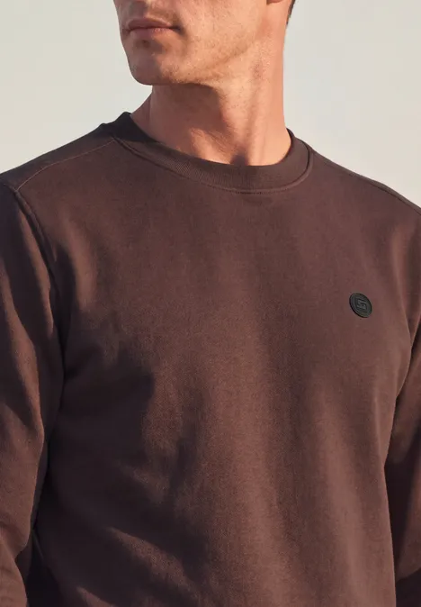 Ein Mann trägt ein schlichtes braunes Sweatshirt mit rundem Logo auf der Brust.