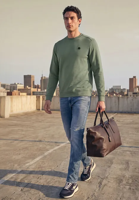 Ein Mann in grünem Pullover und Jeans steht auf einem Dachparkplatz mit Reisetasche und Blick auf die Stadt.