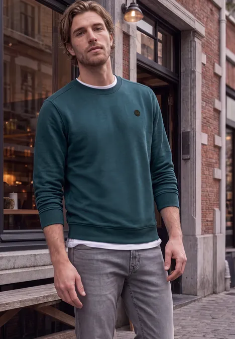 Ein Mann in grünem Pullover und grauen Jeans steht entspannt vor einem Laden auf Kopfsteinpflaster.